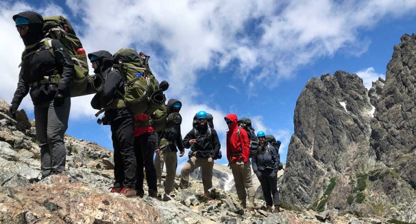 A group of people wearing backpacks make their way over a rocky landscape. There are tall rock formations in the background.